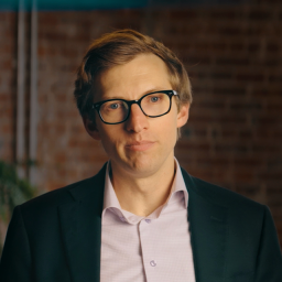 A man with light brown hair and glasses is wearing a dark blazer and light-colored shirt, standing indoors in front of a brick wall.