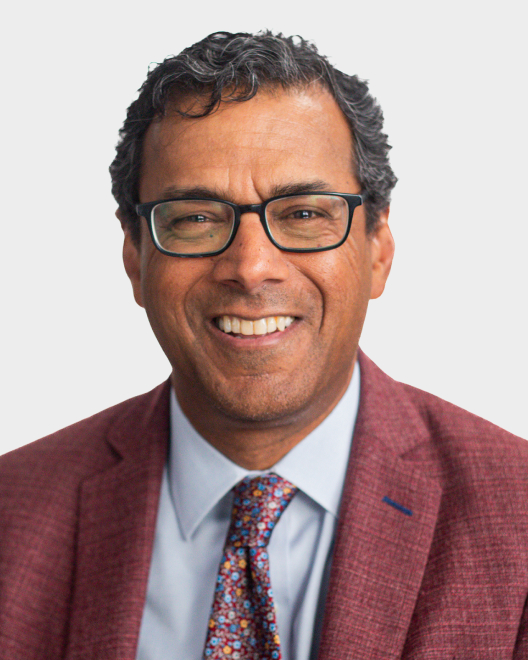 A man wearing glasses, a maroon blazer, a light blue shirt, and a colorful patterned tie, smiling against a plain light background.