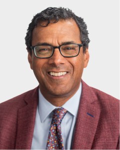A man wearing glasses, a maroon blazer, a light blue shirt, and a colorful patterned tie, smiling against a plain light background.