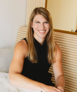 A woman with long blonde hair wearing a sleeveless black top sits at a table, smiling at the camera.