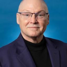A middle-aged man with glasses, short light hair, and a mustache, wearing a dark blazer and black turtleneck, poses in front of a blue background.