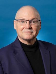 A middle-aged man with glasses, short light hair, and a mustache, wearing a dark blazer and black turtleneck, poses in front of a blue background.