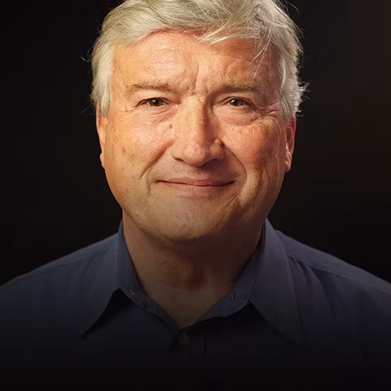 Older man with gray hair and a blue collared shirt smiles slightly, facing forward against a dark background.