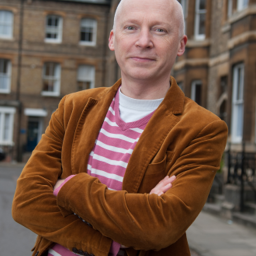 A person with a shaved head wearing a brown corduroy jacket and a pink striped shirt stands outdoors with arms crossed in front of brick buildings.