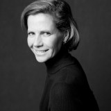 Black and white portrait of a woman with short hair wearing a dark turtleneck, smiling and looking slightly over her shoulder against a plain background.