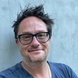 A man with short, messy dark hair, glasses, and stubble smiles slightly at the camera. He is wearing a blue t-shirt and a necklace, standing against a plain light gray background.