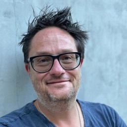 A man with short, messy dark hair, glasses, and stubble smiles slightly at the camera. He is wearing a blue t-shirt and a necklace, standing against a plain light gray background.