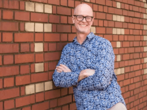 A man with glasses and a patterned blue shirt stands smiling with arms crossed in front of a brick wall.