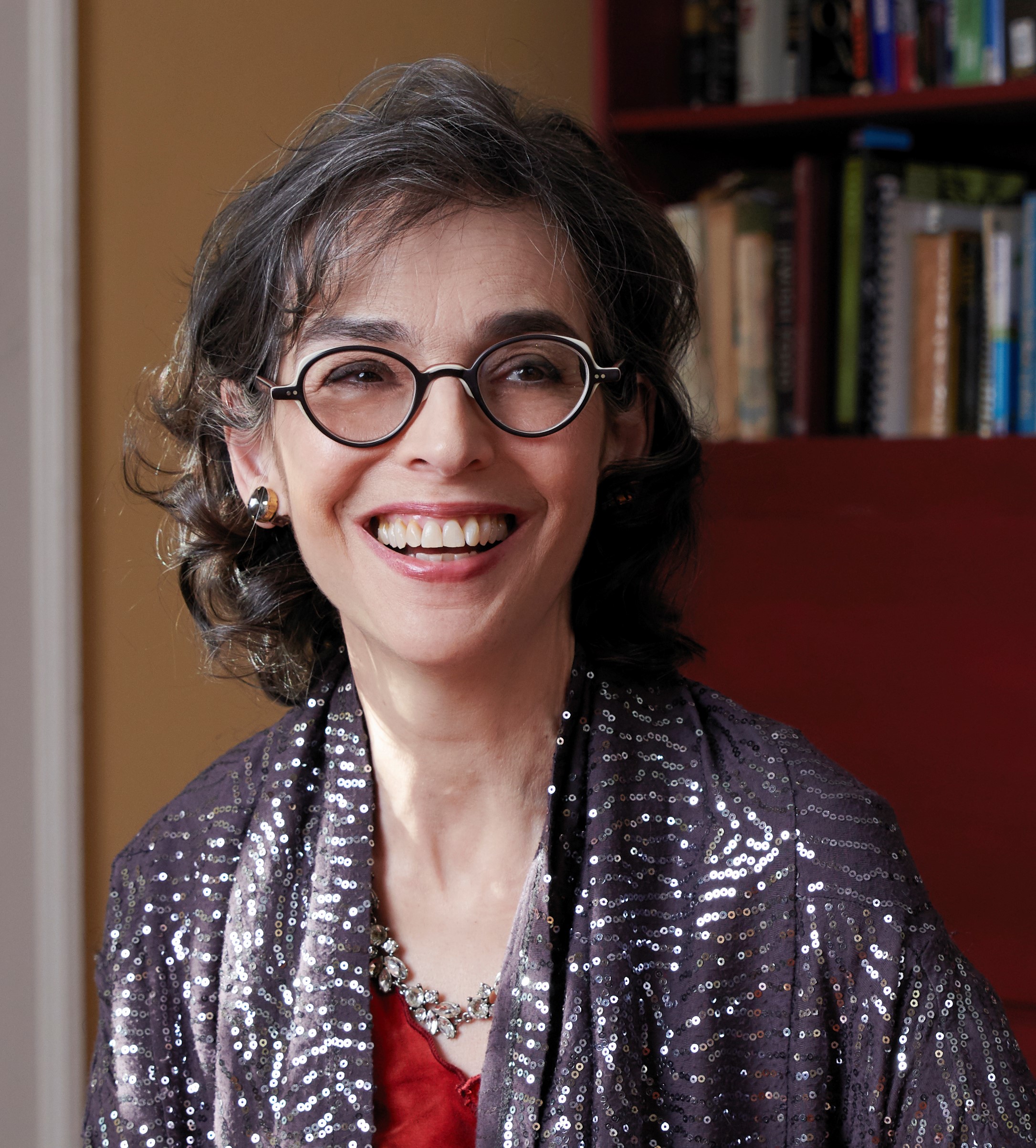 A woman with glasses and curly hair, smiling, wearing a sequined shawl and a red top, seated in a room with a bookshelf in the background.