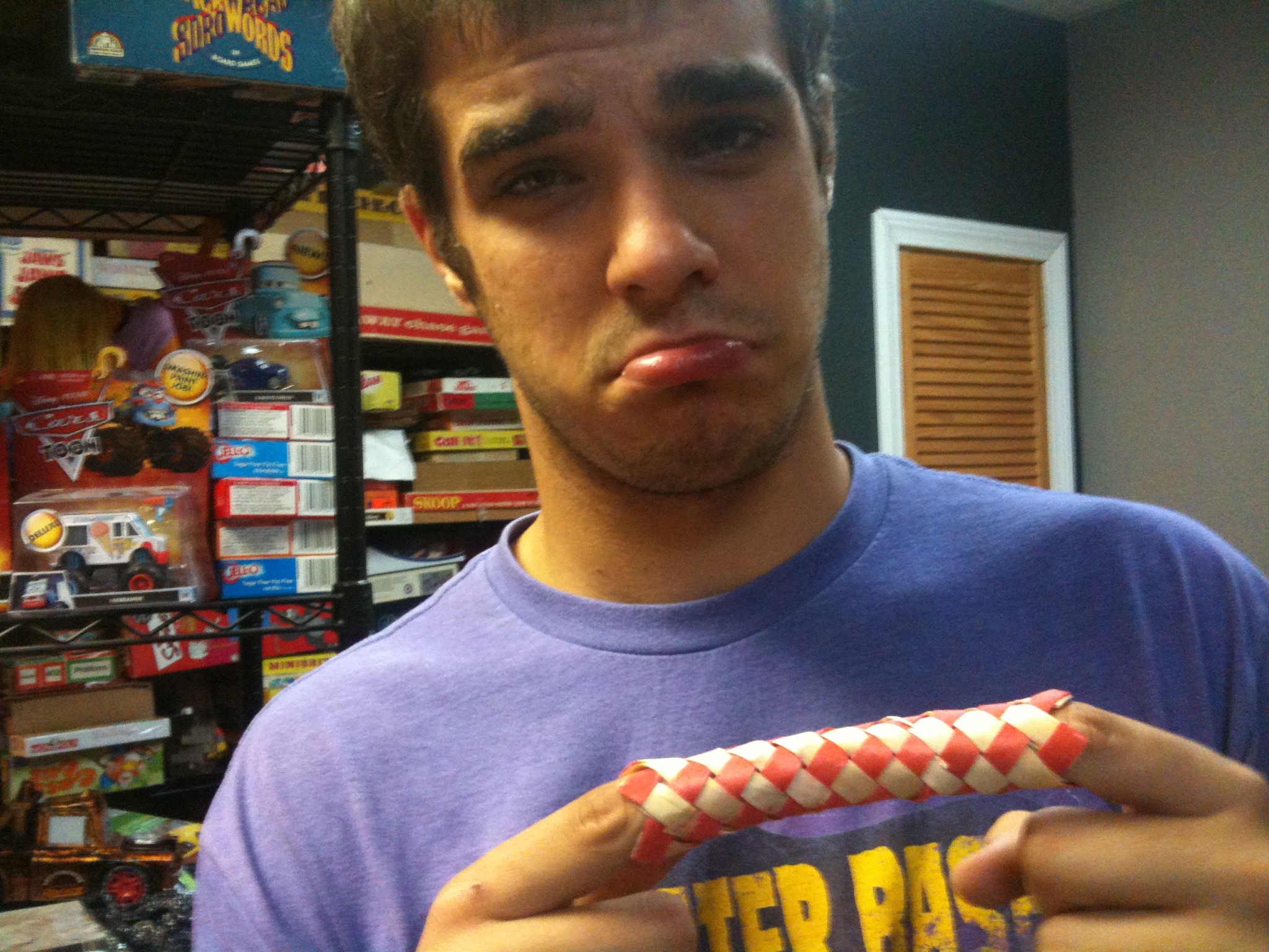A person with a sad expression holds their fingers trapped in a red and white Chinese finger trap, with shelves of toys in the background.