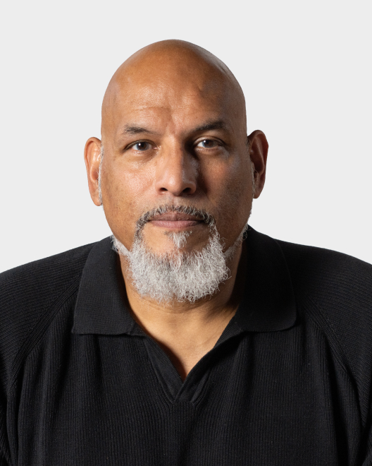 Bald man with a gray goatee wearing a black collared shirt, looking directly at the camera against a plain light background.