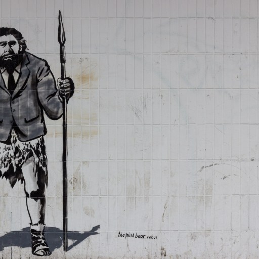 A Neanderthal man holding a briefcase in one hand and a spear in the other.
