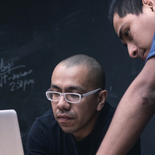 Two men look at a laptop in front of a blackboard.