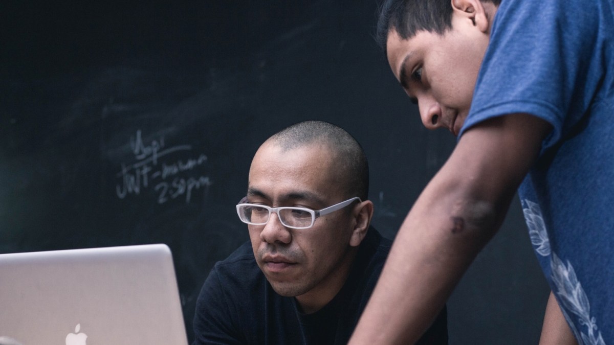 Two men look at a laptop in front of a blackboard.