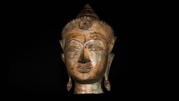 A bronze Buddha head sculpture with ornate crown and elongated earlobes, set against a black background, embodies the philosophies of self through its serene expression and timeless artistry.