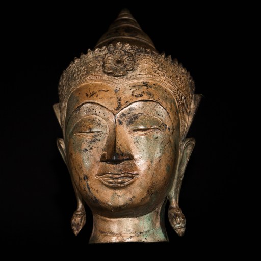 A bronze Buddha head sculpture with ornate crown and elongated earlobes, set against a black background, embodies the philosophies of self through its serene expression and timeless artistry.