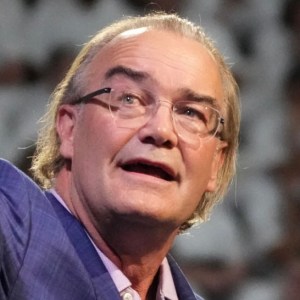 An older man with glasses and gray hair, wearing a blue suit jacket, is speaking. He looks slightly upwards, with a background of blurry, out-of-focus shapes.