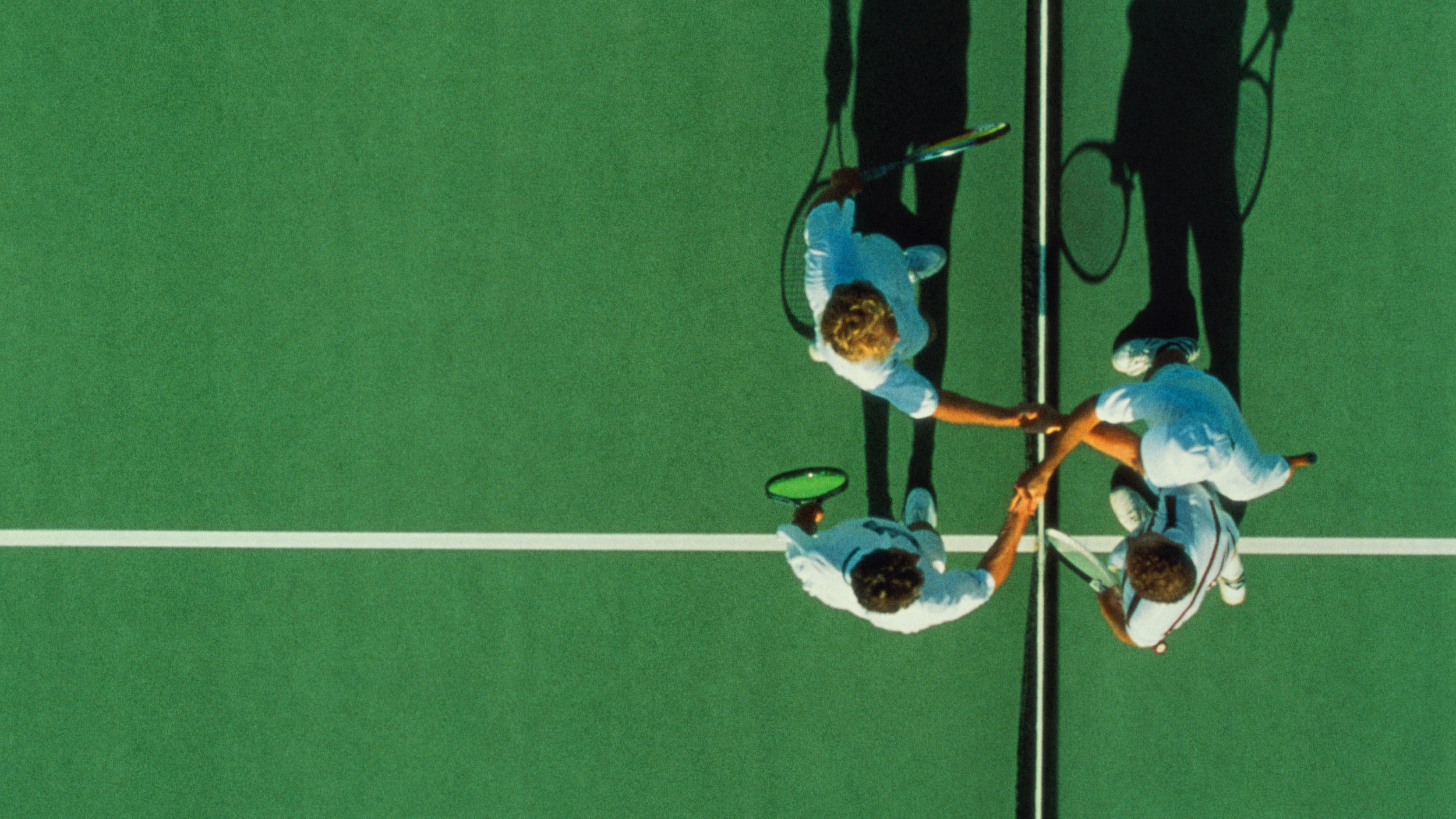 An aerial view captures two pairs of tennis players, akin to The Four Enemies, shaking hands at the net on a lush green court.