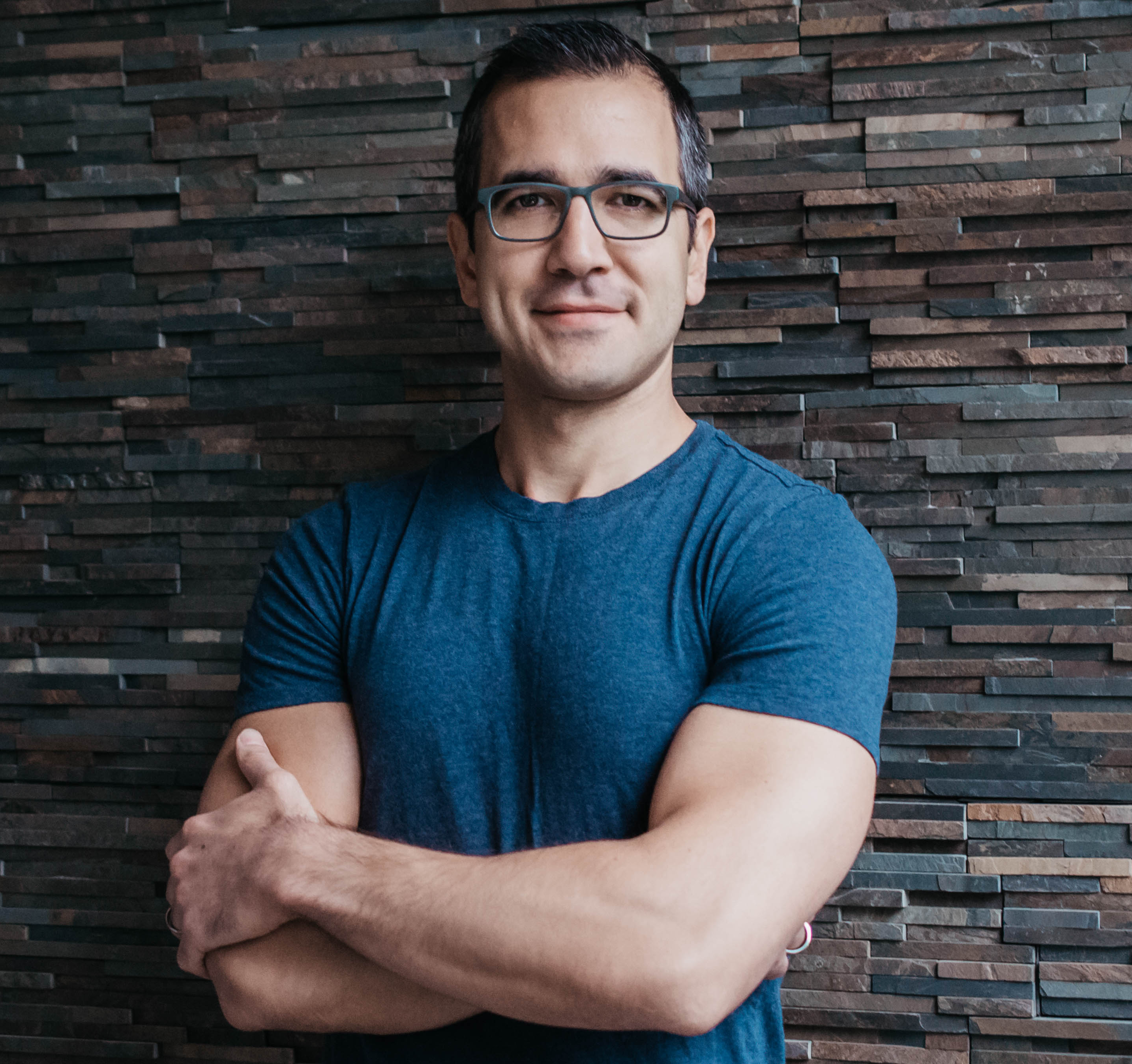 a man wearing glasses standing in front of a brick wall.