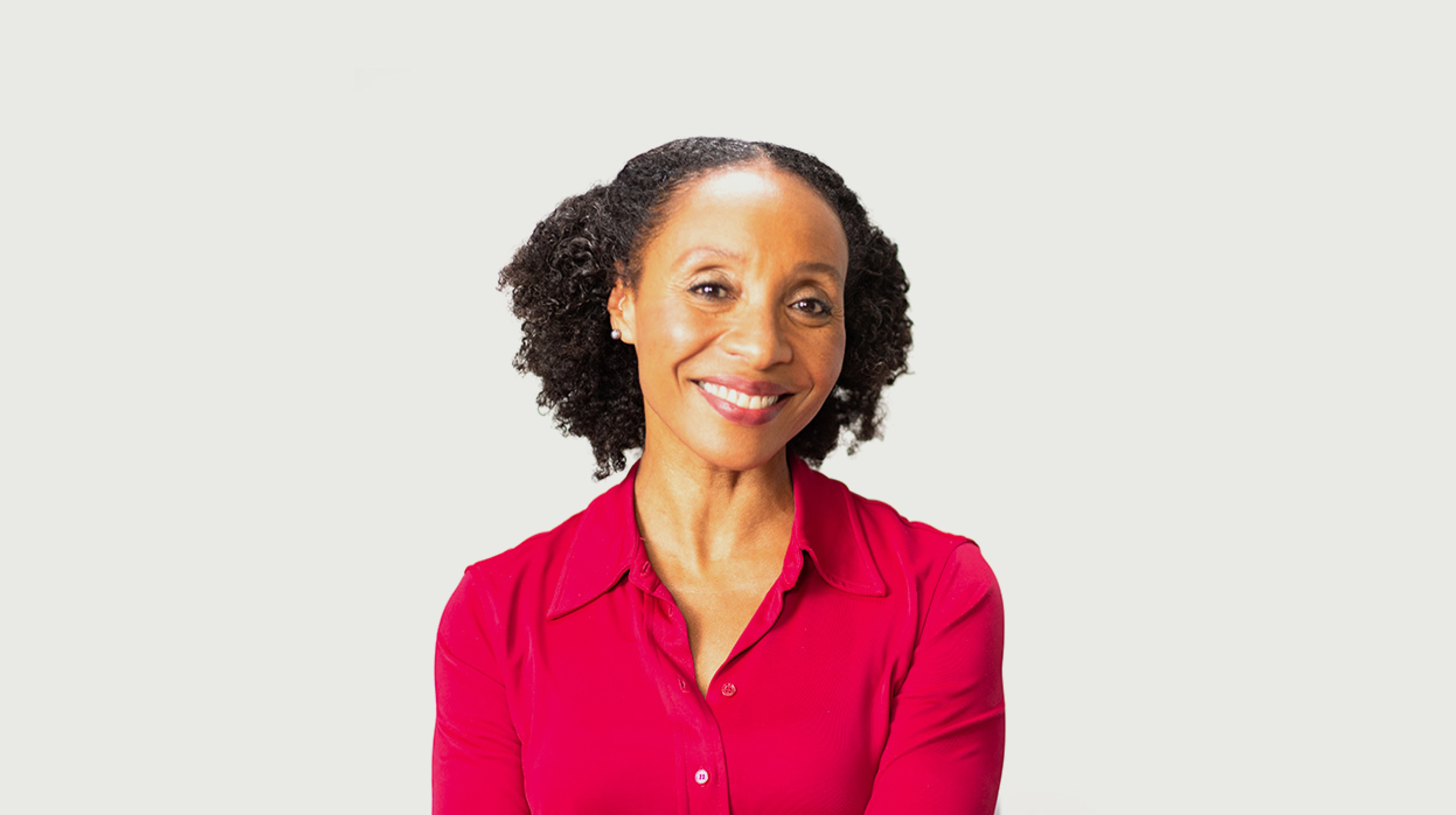 A woman with curly hair, wearing a red blouse, smiles at the camera against a plain white background.