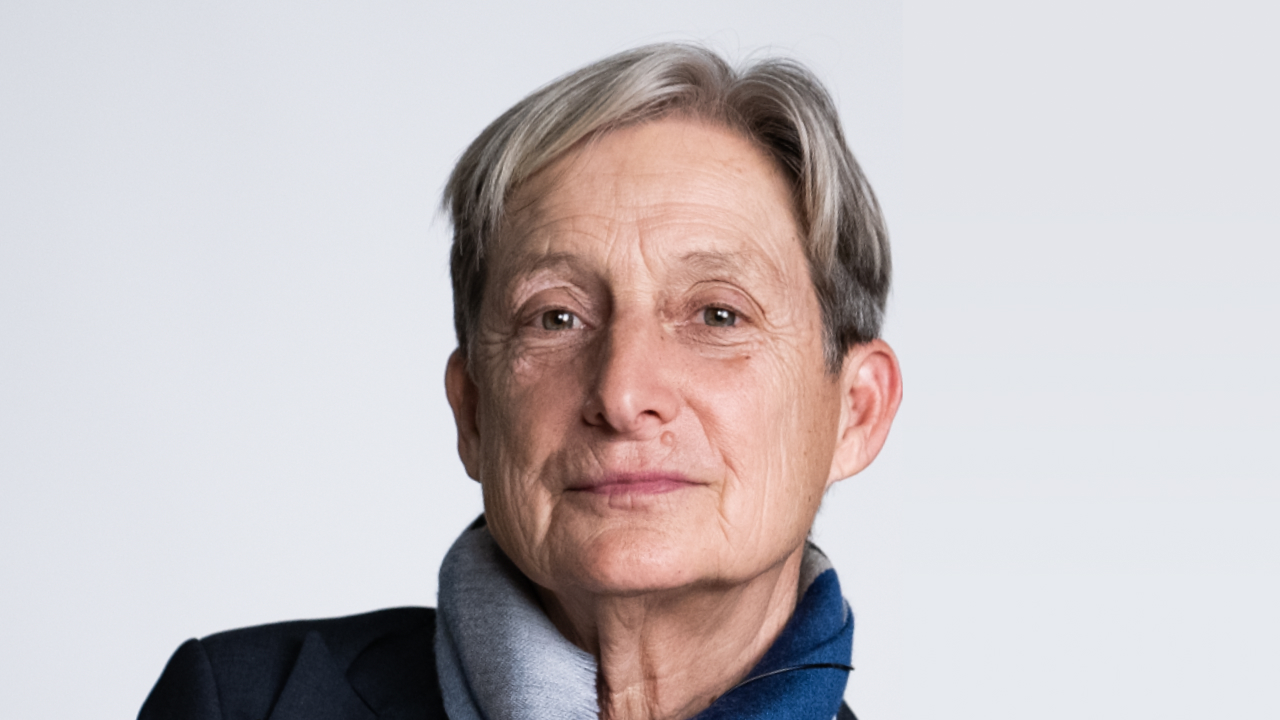 A person with short gray hair, wearing a dark jacket and a gray-blue scarf, looks at the camera against a plain light background.