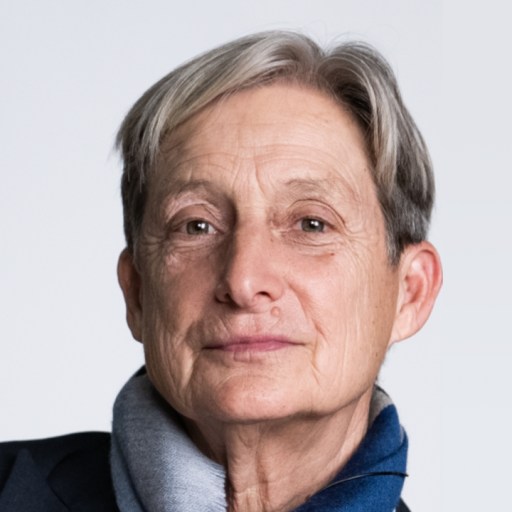 A person with short gray hair, wearing a dark jacket and a gray-blue scarf, looks at the camera against a plain light background.