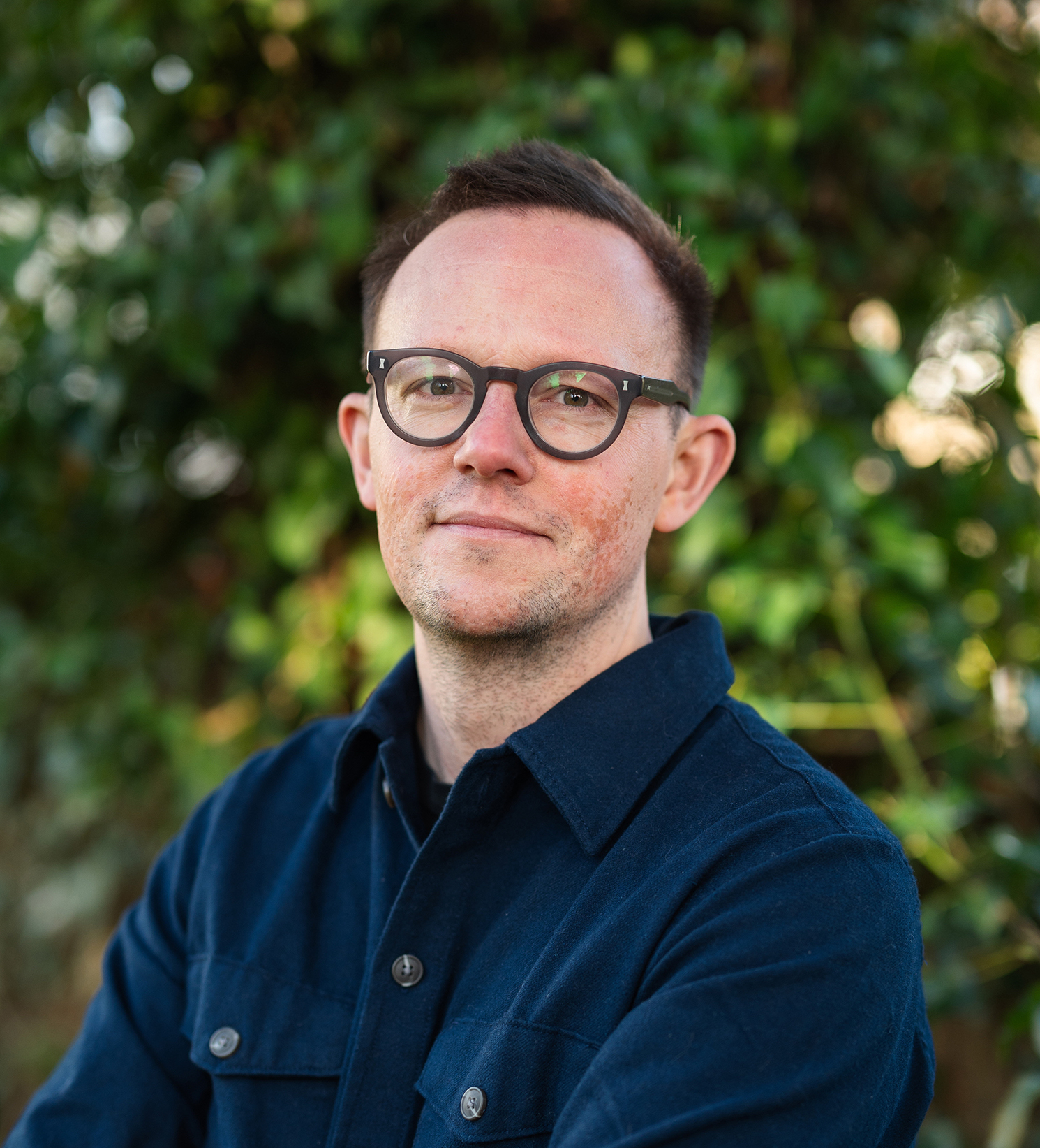 A man in glasses standing in front of bushes.