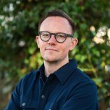 A man in glasses standing in front of bushes.