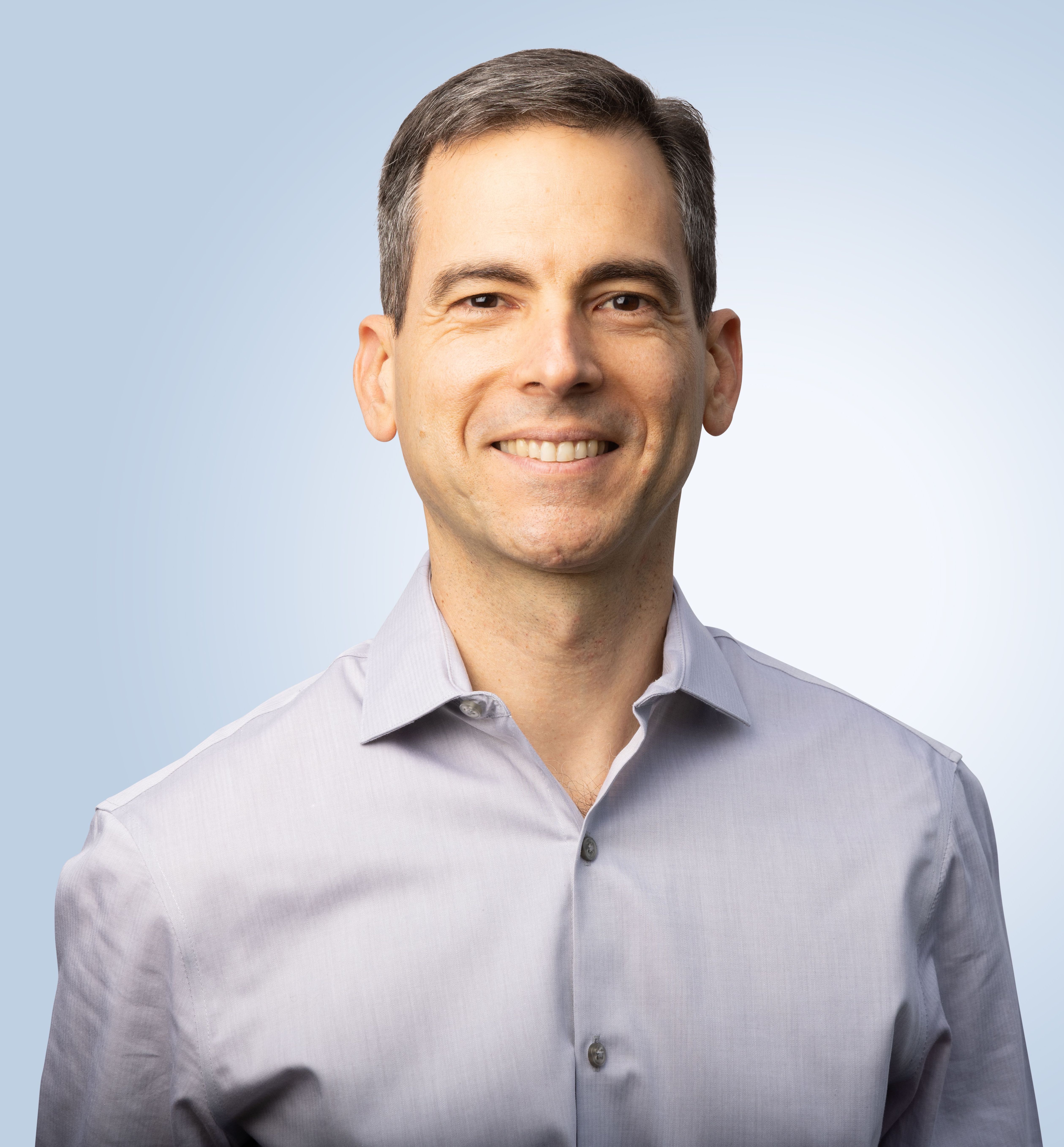 A man in a gray shirt smiling in front of a blue background.