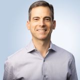 A man in a gray shirt smiling in front of a blue background.