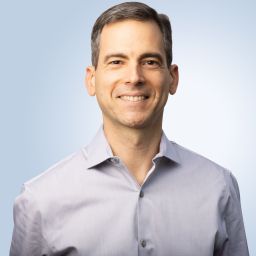 A man in a gray shirt smiling in front of a blue background.