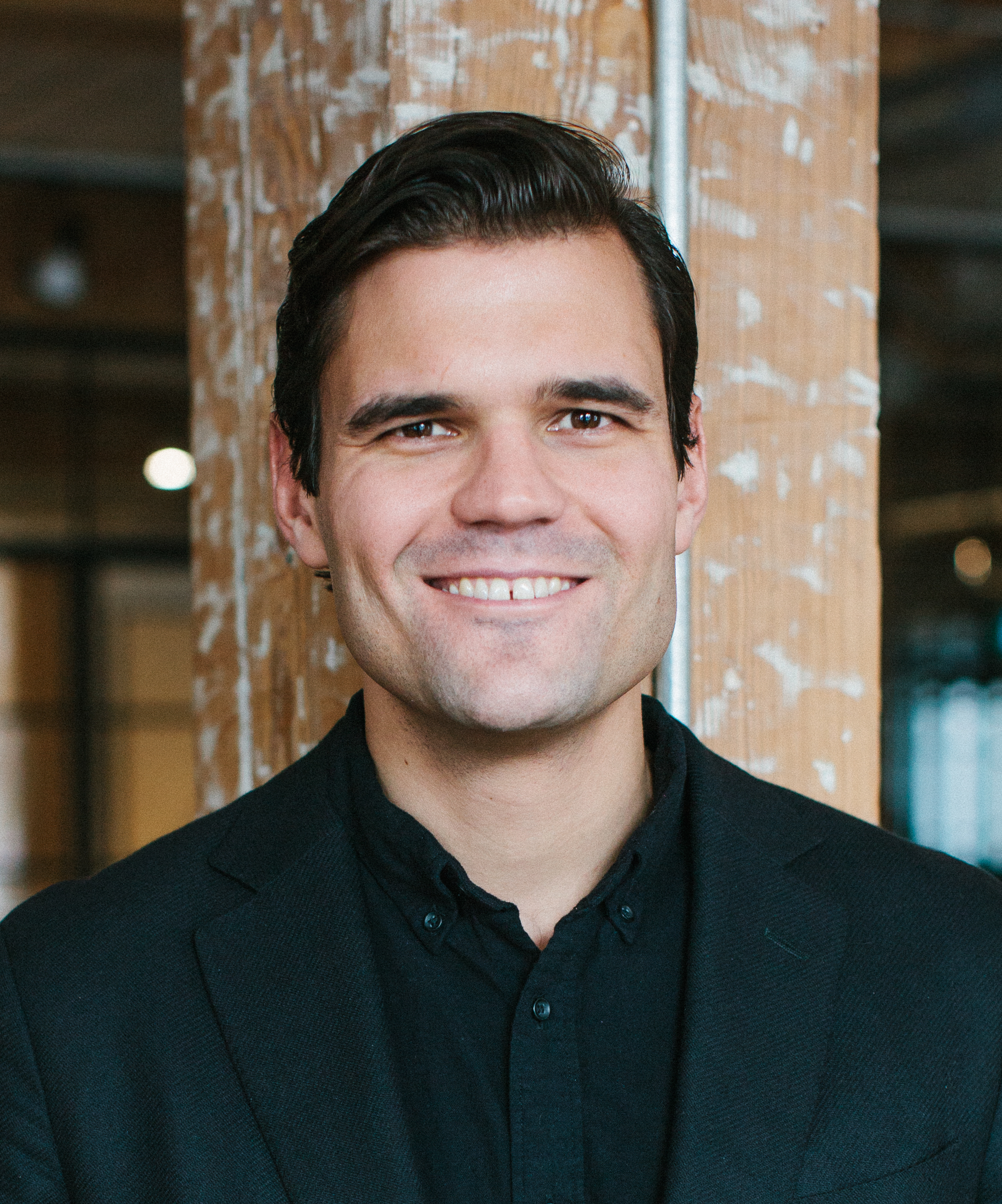 A man in a black shirt smiling in front of a wooden wall.
