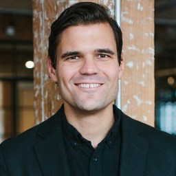 A man in a black shirt smiling in front of a wooden wall.