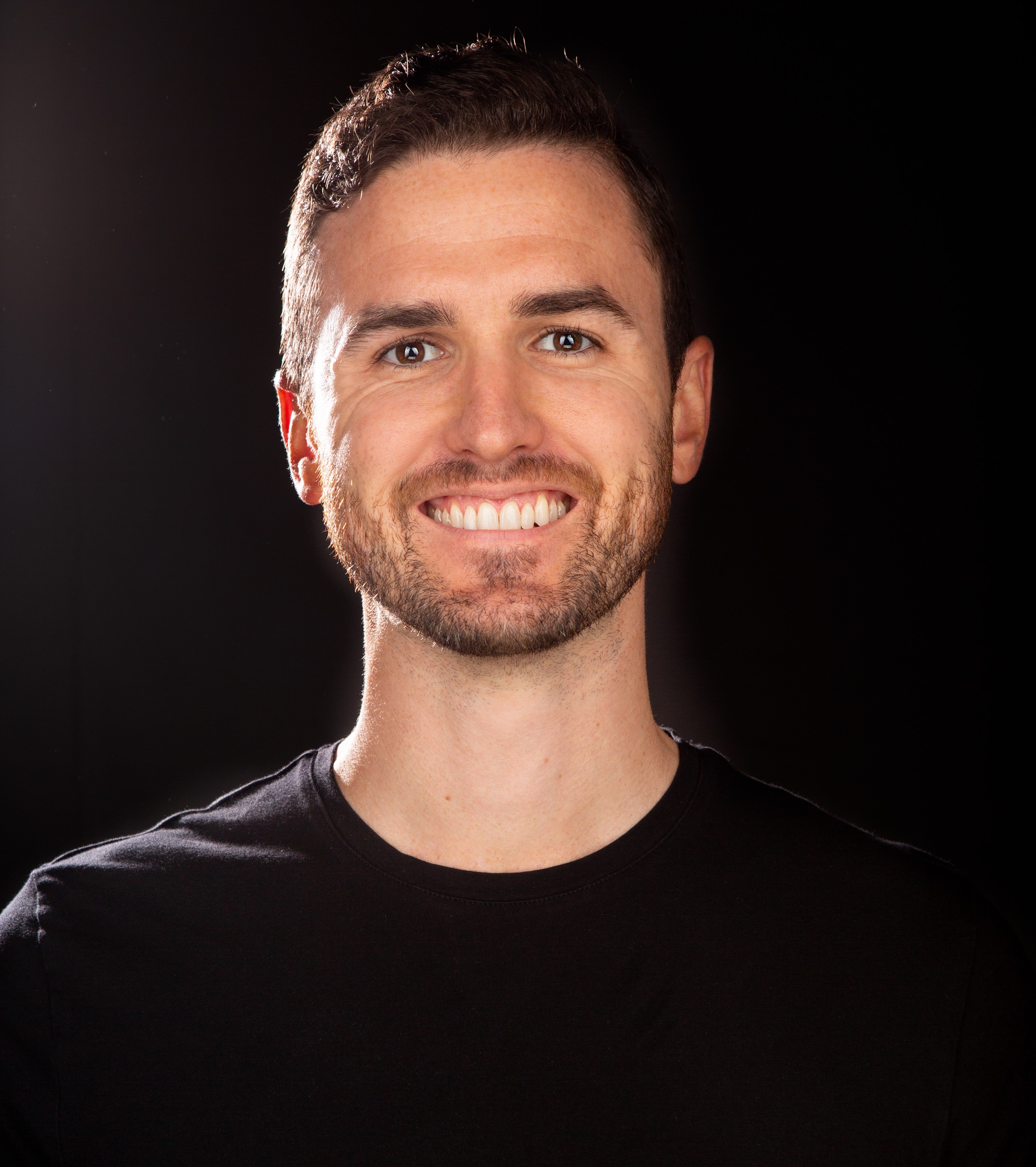 A man in a black shirt smiling in front of a black background.