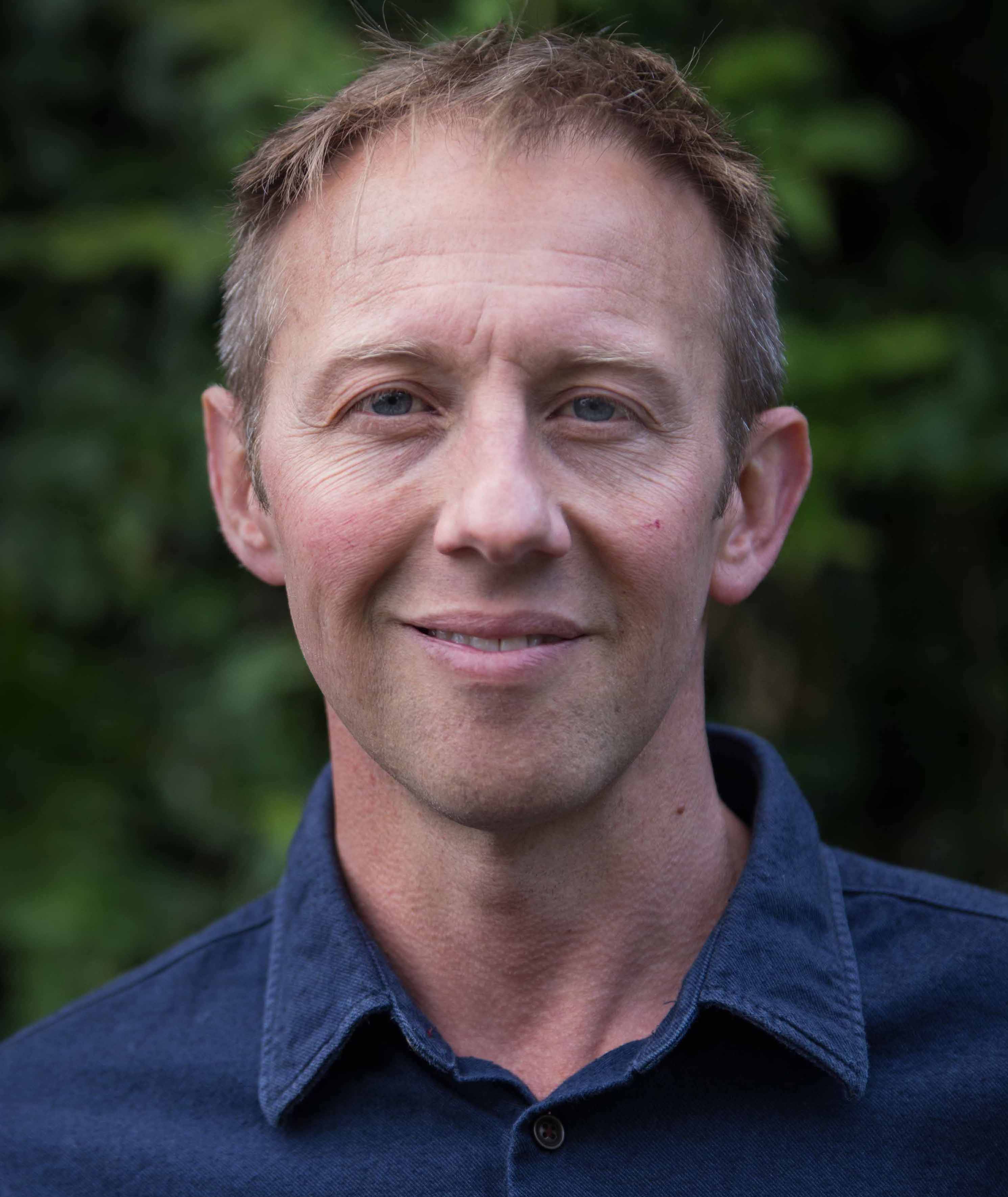 A man in a blue shirt smiling in front of trees.
