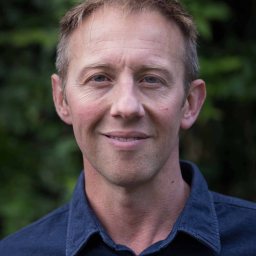 A man in a blue shirt smiling in front of trees.