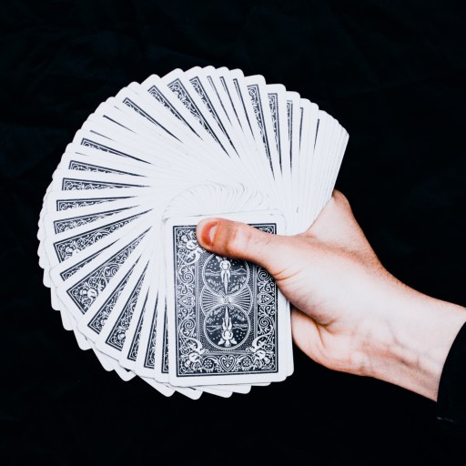 A hand holds and fans out a deck of playing cards with patterned backs against a black background.