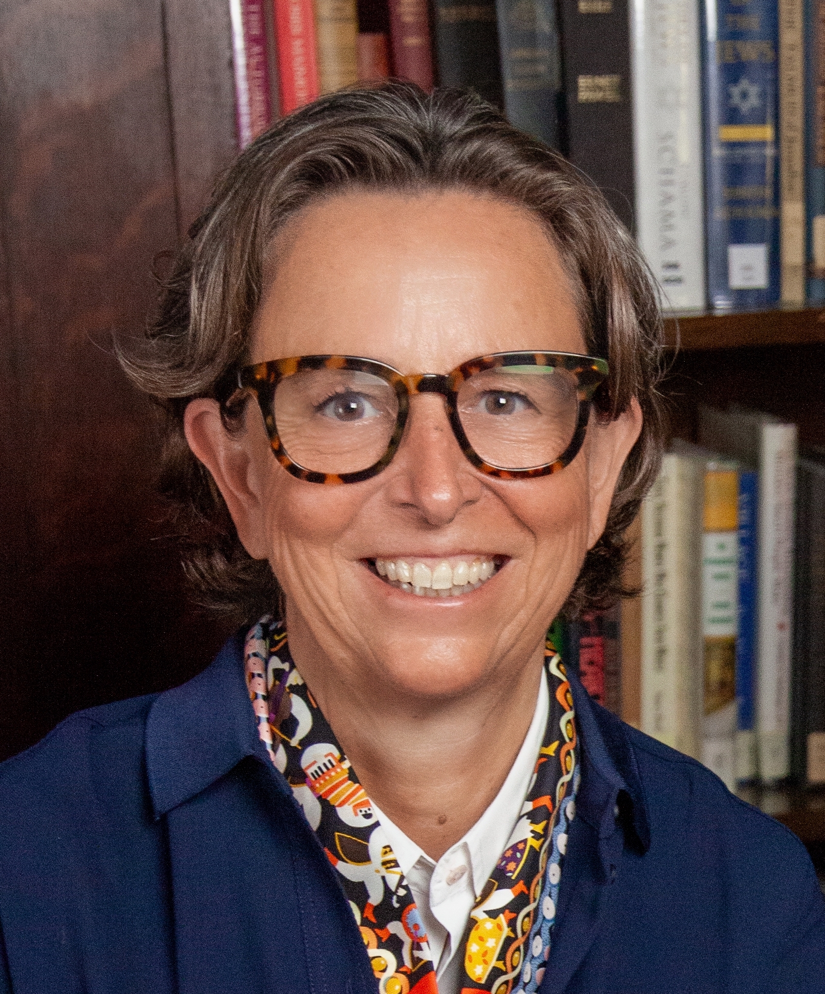 A woman in glasses is smiling in front of a bookcase.