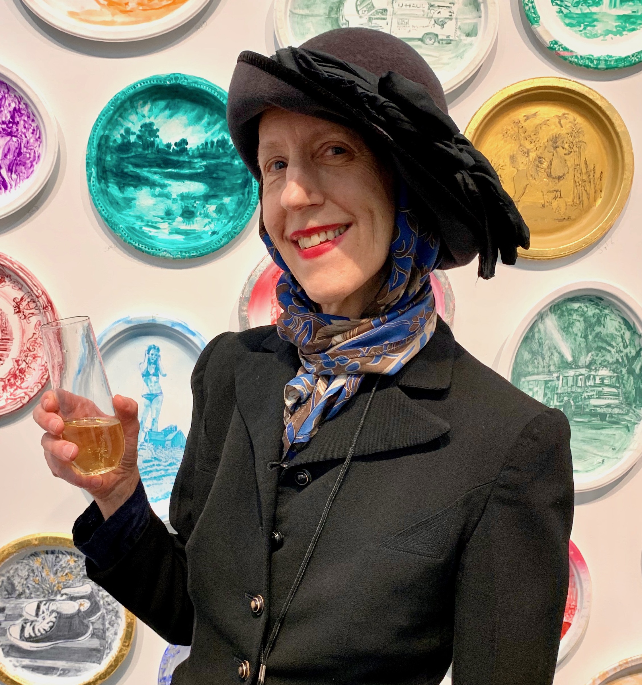 A woman holding a glass of wine in front of a wall of plates.