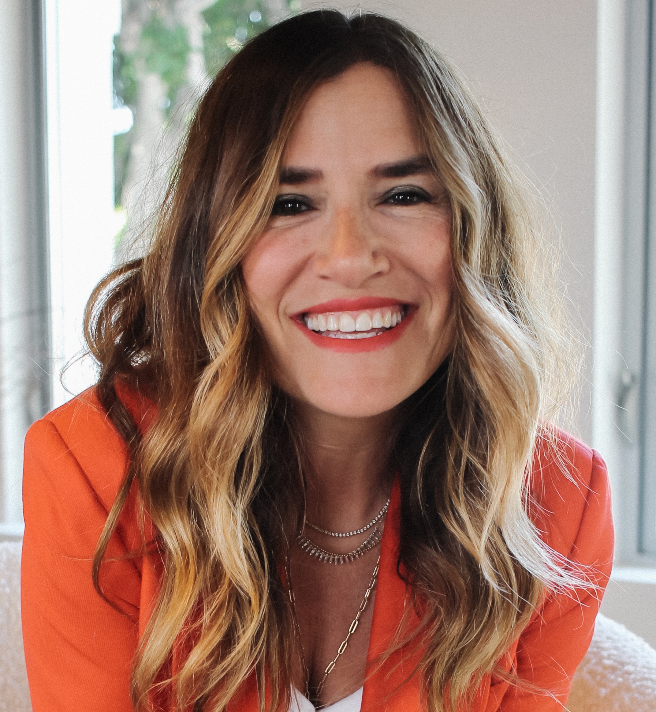 A smiling woman in an orange blazer sitting on a couch.