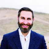 A man with a beard smiling in front of a mountain.