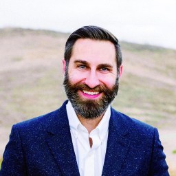A man with a beard smiling in front of a mountain.