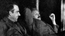 Two men sit closely together, one smiling and the other reclining with a relaxed posture against a dark background.