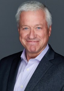 A man in a suit and a purple shirt is smiling.