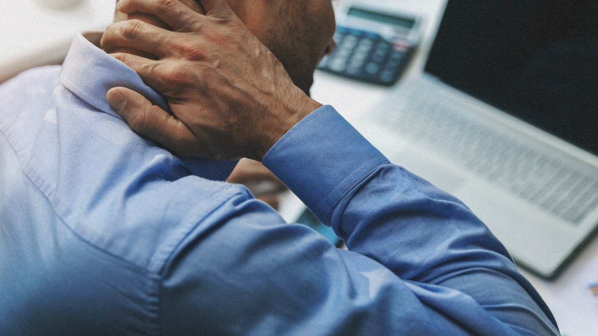 A man in a blue shirt is holding his neck in front of a laptop, possibly experiencing inflammation.