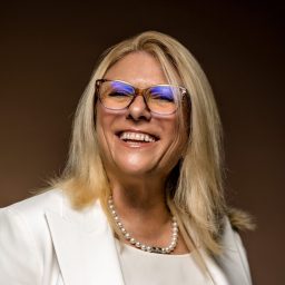 A smiling woman in glasses wearing a white blazer and pearls.