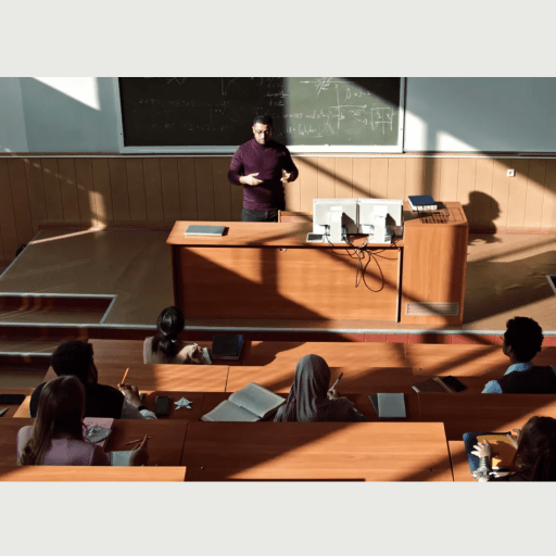 Lecturer standing in front of a classroom, teaching college admissions students seated at desks with sunlight casting shadows.