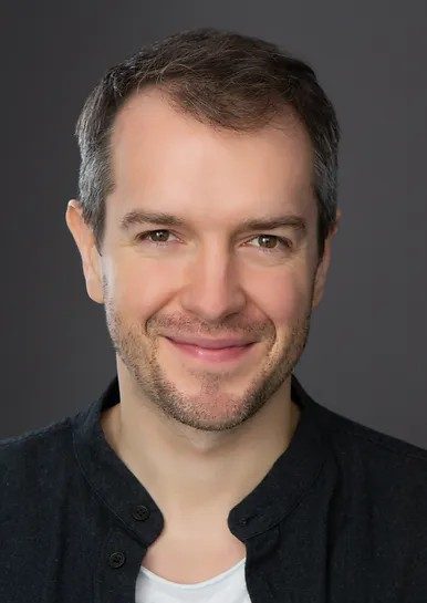 A headshot of a smiling man with stubble wearing a dark-colored shirt over a white t-shirt against a gray background.