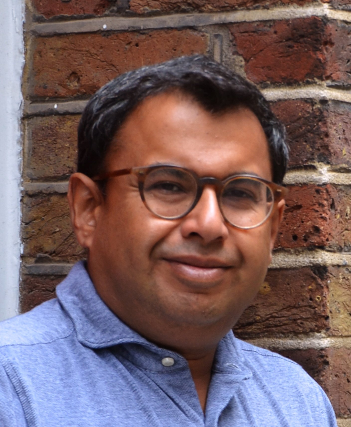 Man with glasses smiling against a brick wall background.