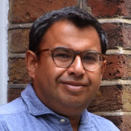 Man with glasses smiling against a brick wall background.